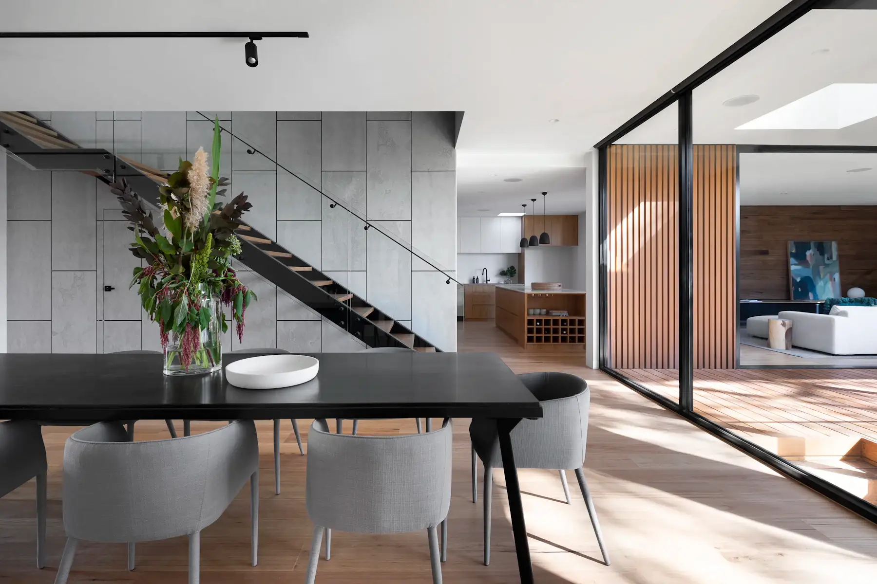 Inspiration image: open-plan dining area with a black dining table, concrete-look wall panels, floating steel staircase, timber slat partition and glass sliding doors