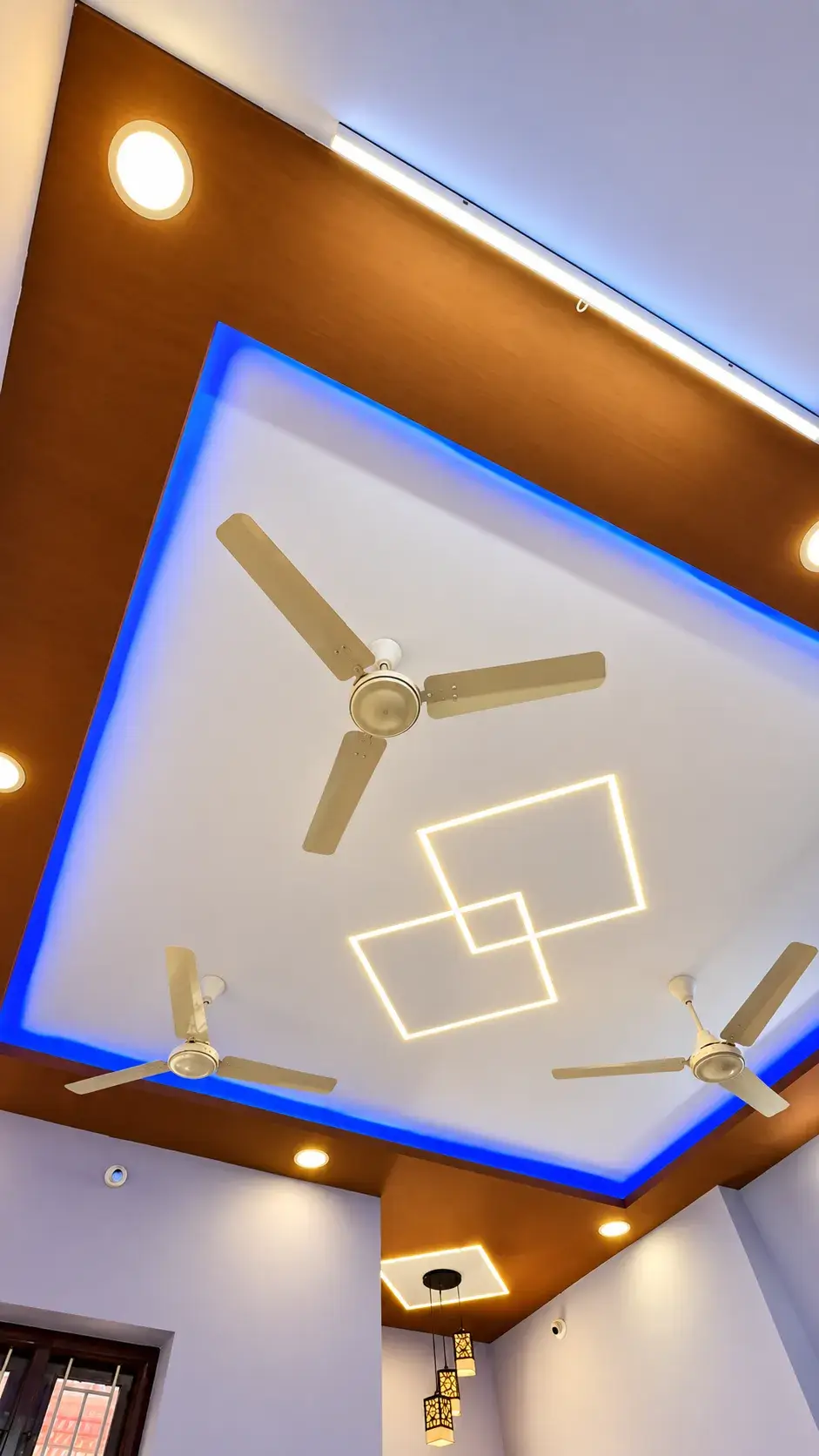 Layered false ceiling with a wood-finish border, blue cove lighting, overlapping square light profiles and pendant lanterns